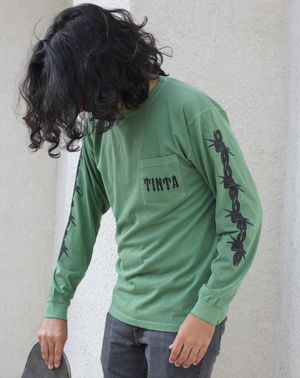 green long sleeve shirt with barbed wire on sleeves and the word "tinta" on the front pocket. Man holding skateboard with shoulder length curly hair.